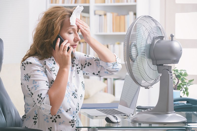 AC_B_018-GettyImages-1198411657 When Do I Replace My Air Conditioner? Woman on the phone attempting to cool herself with a fan.