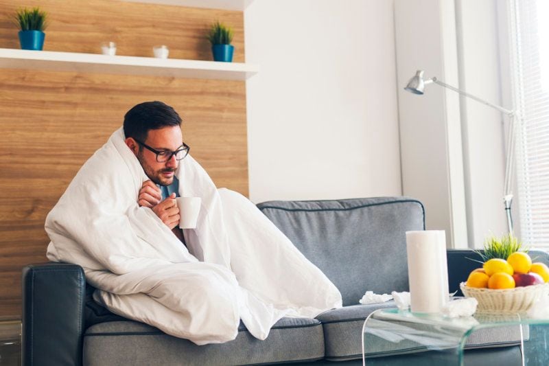 Why Is My Furnace Blowing Cold Air? Image shows a man wrapped in a white blanket and sitting on a grey couch looking cold.|