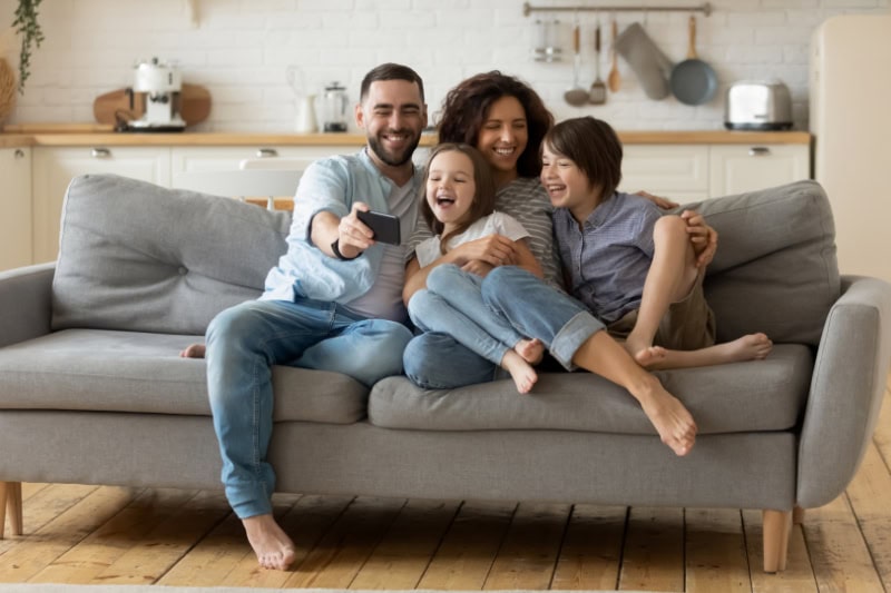 What Are the Signs that I Need a New Furnace? Smiling parents with little kids laughing using smartphone together sitting on couch at home. Happy father holding phone taking selfie with children. Family watching video having fun with cellphone.
