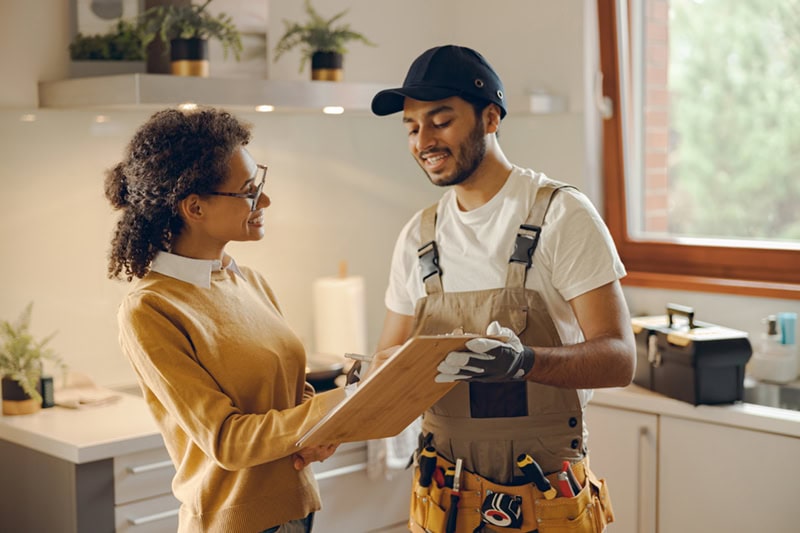 A smiling service technician in overalls and a tool belt reviewing paperwork on a wooden clipboard with a woman inside a bright kitchen.