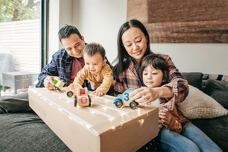 A family sitting on a sofa and playing together with toy cars on top of a cardboard box lined with fairy lights.