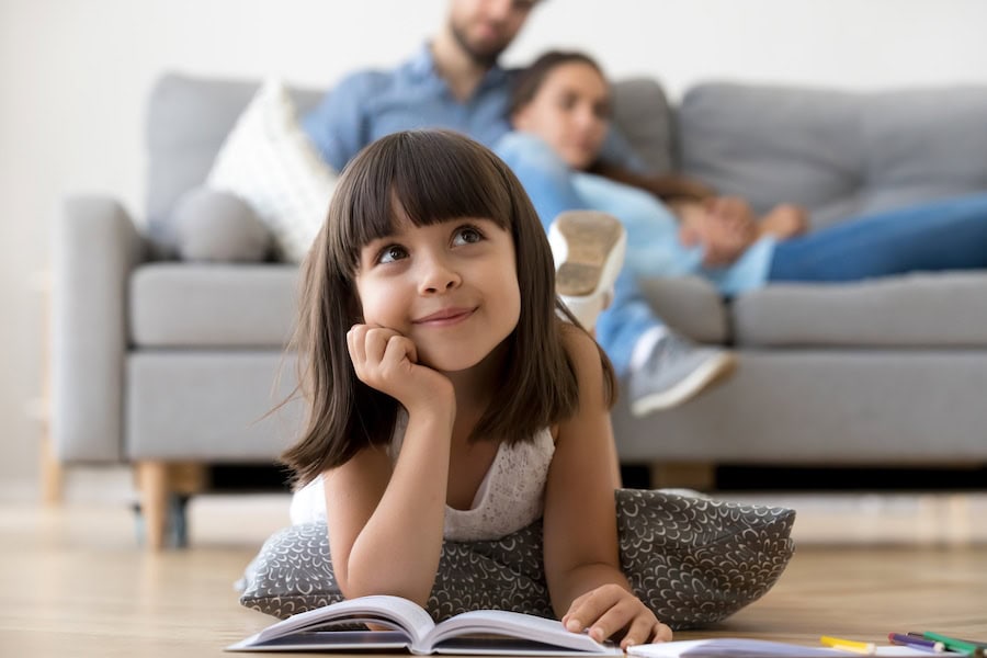 Close up little adorable thoughtful smiling daughter dreaming lying at cushion on warm floor with book in living room at modern home